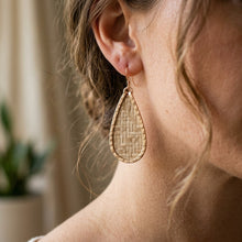 Close-up of a person wearing a gold woven teardrop earring with a blurred indoor background.