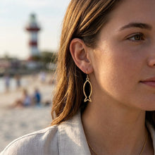 Woman wearing gold fish-shaped earrings with a beach and lighthouse in the background