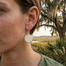 Close-up of a person wearing a shell-shaped earring with a natural landscape in the background.