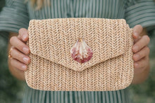 Person holding a woven clutch with a shell decoration against a blurred background