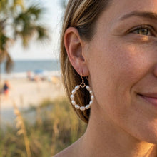 Close-up of a woman wearing pearl hoop earrings with a beach in the background