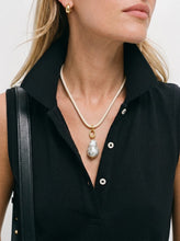 A close-up of a model wearing a gold-tone necklace with a large, irregular white pearl pendant, styled with a black sleeveless top and a black shoulder bag strap.