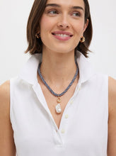 A woman wearing a white sleeveless collared top and a layered necklace with a navy and white striped cord, a gold ring, and a white baroque pearl pendant.
