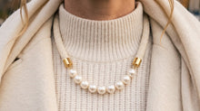 A close-up of a person wearing a cream-colored knit turtleneck sweater and a cream cord necklace with white pearls and gold-toned metal accents.